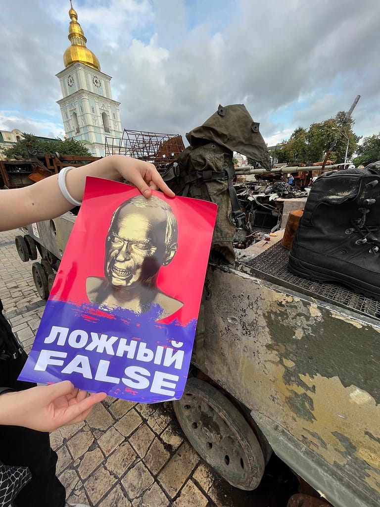 Protest poster held next to a burned Russian military vehicle